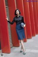 A woman leaning against a red wall wearing a black shirt and blue skirt.