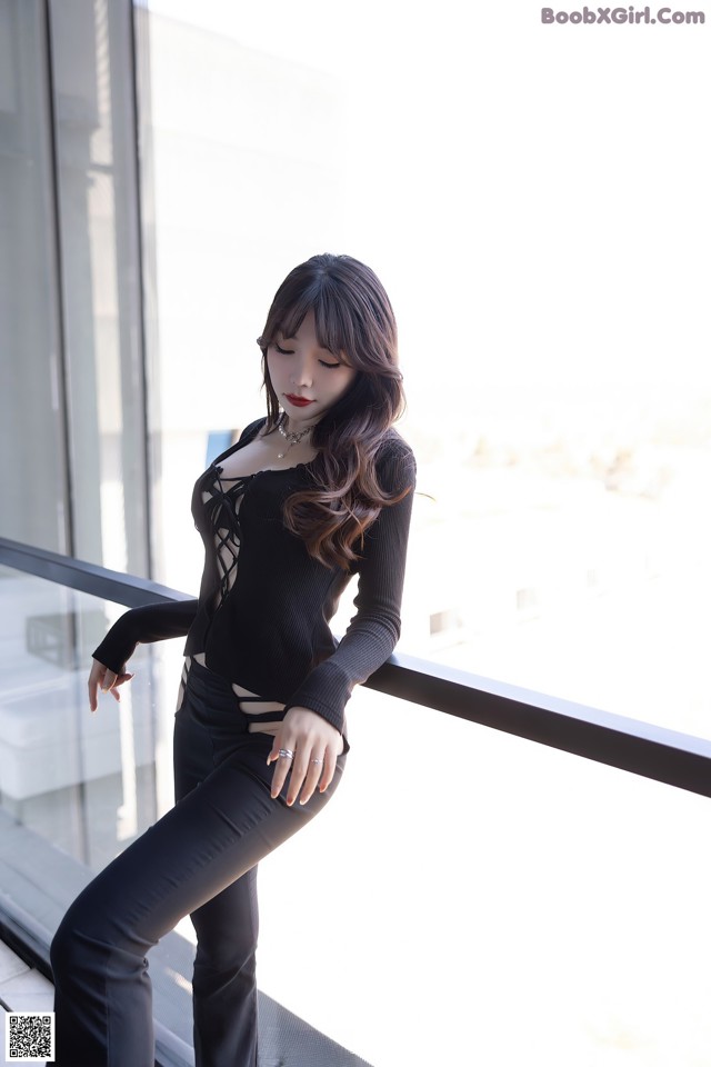 A woman in a black top and black pants posing by a window.