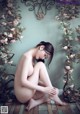 A naked woman sitting on the floor in front of a wall of flowers.